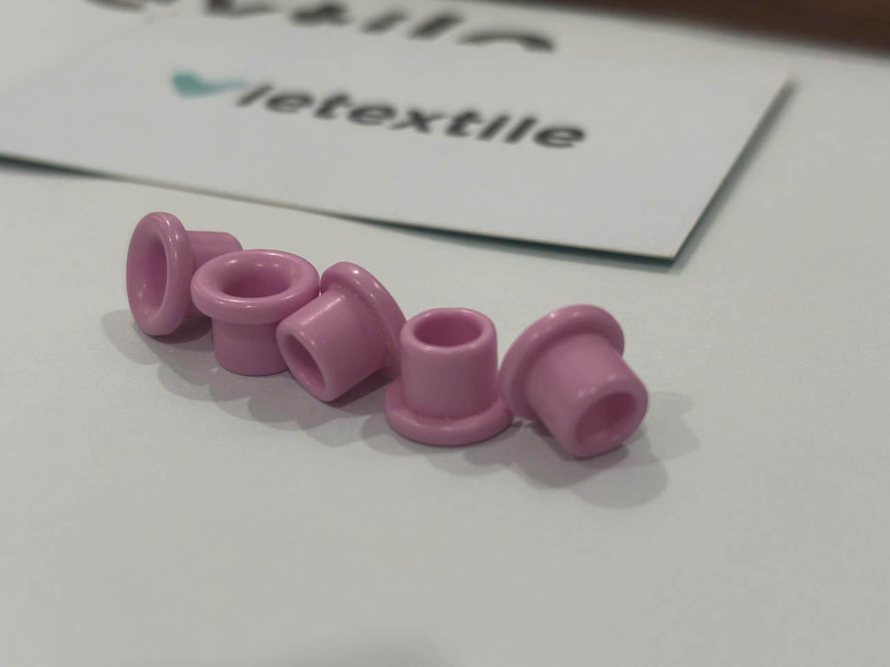 Ceramic Eyelet Risks: 7 Preventive Checks 3 Five pink ceramic eyelet risk set against a white background, with a Vietextile business card displaying contact information positioned behind them.